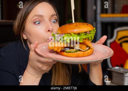 Porträt einer überraschten Frau, die mit breiten Augen auf einen Burger starrt, mit Salat, Huhn, Zwiebeln auf einem großen Teller im Café-Restaurant. Stockfoto