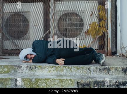 Gefrorener Obdachloser schläft auf Beton in der Nähe des Lüftungssystems des Gebäudes. Zur Frage, wie Obdachlose die Winterkälte überleben. Stockfoto