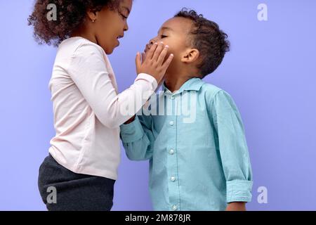 Zwei fröhliche schwarze amerikanische Geschwister, Schwester küsst ihren Bruder, isoliert auf lila Hintergrund. Porträt süßer Kinder in lässiger Kleidung. Junge und Mädchen Stockfoto