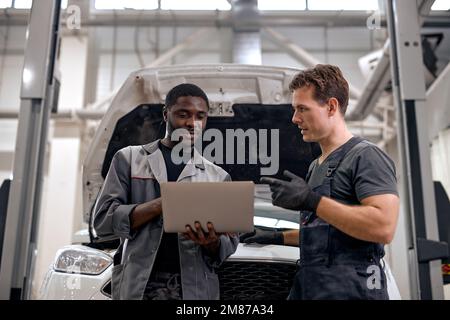 Black Car Mechanic und kaukasischen Werkstatt Überprüfung der Diagnoseergebnisse auf Laptop, Computer. Erläutert eine Motorabbruchanalyse für Mechanic. Car Service-Mitarbeiter Stockfoto