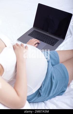 Schwangere Frauen saßen auf dem Bett und benutzten Laptops Stockfoto