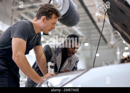 Schwarze und amerikanische oder kaukasische Männer können alles reparieren. Gutaussehende Automechaniker in Overalls Uniform Überprüfung des Motors unter der Haube in modernen sauberen Auto se Stockfoto