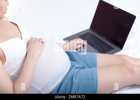 Schwangere Frauen saßen auf dem Bett und benutzten Laptops Stockfoto