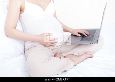 Schwangere Frauen saßen auf dem Bett und benutzten Laptops Stockfoto