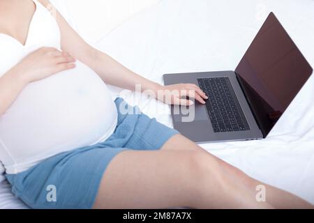 Schwangere Frauen saßen auf dem Bett und benutzten Laptops Stockfoto