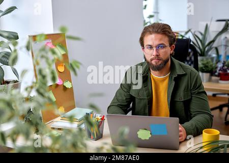 Porträt eines selbstbewussten, weißen Designers, der allein in einem modernen, hellen Büro arbeitet, lässig gekleidet. Ein gutaussehender, bärtiger Kerl, der sich auf Wor konzentriert hat Stockfoto