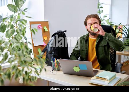 Ein bärtiger Kerl arbeitet mit einem Laptop im Büro und trinkt Kaffee, allein, Erfolg und Geschäftskonzept. Hübscher, netter Mann in lässigem Outfit konzentriert sich auf die Arbeit Stockfoto