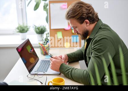 Der junge Mann sitzt mit dem Smartphone und ruht sich während der Arbeit in einem hellen, modernen Büro aus, allein. Ein bärtiger weißer Kerl in Freizeitkleidung macht eine Pause beim Tippen von Messa Stockfoto