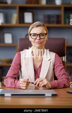 Porträt einer schönen blondie Frau, die die Kamera anschaut und lächelt. Erwachsene stilvolle selbstbewusste Geschäftsfrau trägt Brillen sitzen am Arbeitsplatz in Stockfoto