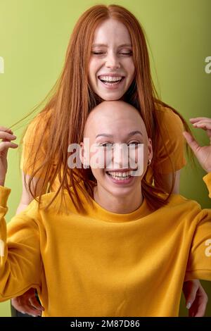 Zwei fröhliche junge kahlköpfige und rothaarige europäische Weiße, die sich in einem Studio-Porträt mit grünem Hintergrund umarmen. Menschen, menschliche Emotio Stockfoto