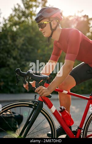 Schneller werden. Selbstbewusster junger Kaukasier passt männlicher Rennfahrer in Sportbekleidung und Schutzhelm sieht konzentriert aus, während er Fahrrad fährt und in der Natur fährt Stockfoto