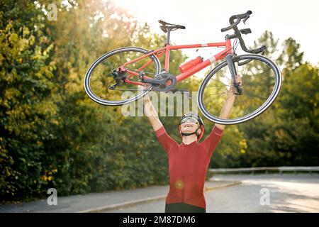 Happy Sporty Männchen in sportlicher Kleidung, Schutzhelm und Brille trägt das Fahrrad nach erfolgreichem Wettkampf und posiert vor der Kamera mit Rot Stockfoto