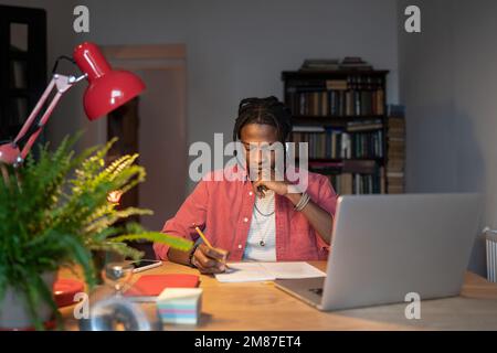 Verwirrter junger schwarzer Mann macht Notizen mit dem Kinn an schwieriger Forschungsarbeit Laptop am Schreibtisch verwenden. Stockfoto