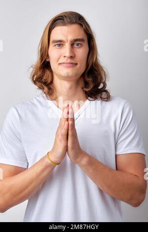 Angenehmer weißer Mann in lässigem T-Shirt, der bettelt und die Hände im Gebet hält, isoliert auf einem hellgrauen Studioporträt. Modellkopien Stockfoto