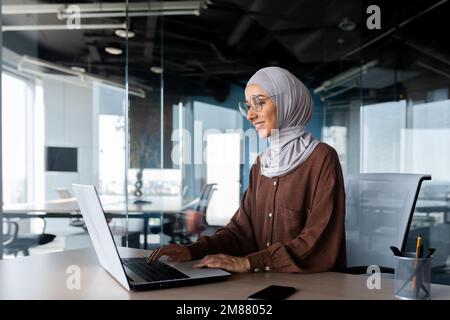 Junge, wunderschöne muslimische Frau im Hijab, die in einem modernen Büro arbeitet, Geschäftsfrau, die ein Notebook bei der Arbeit verwendet, Tastatur eingibt, während sie am Arbeitsplatz sitzt und lächelt und mit dem Erfolg zufrieden ist. Stockfoto