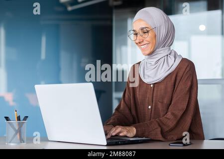 Junge, wunderschöne muslimische Frau im Hijab, die in einem modernen Büro arbeitet, Geschäftsfrau, die ein Notebook bei der Arbeit verwendet, Tastatur eingibt, während sie am Arbeitsplatz sitzt und lächelt und mit dem Erfolg zufrieden ist. Stockfoto