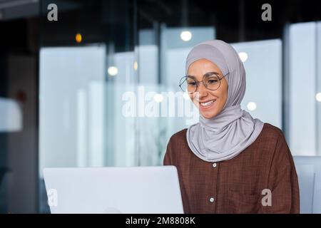 Junge, wunderschöne muslimische Frau im Hijab, die in einem modernen Büro arbeitet, Geschäftsfrau, die ein Notebook bei der Arbeit verwendet, Tastatur eingibt, während sie am Arbeitsplatz sitzt und lächelt und mit dem Erfolg zufrieden ist. Stockfoto