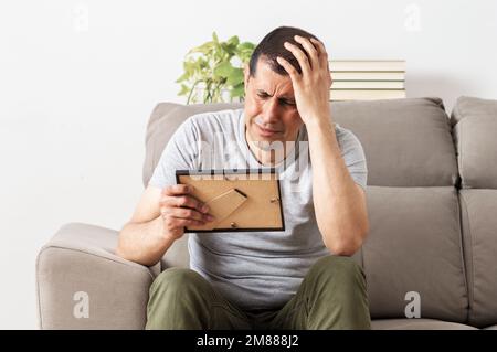 Trauriger Ehemann, der seine Frau nach der Scheidung vermisst und sich ein Bild von dem Paar ansieht, das im Wohnzimmer in einem Haus auf der Couch sitzt Stockfoto
