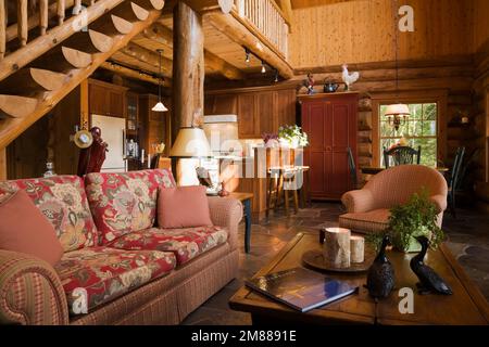 Gepolstertes Sofa, Sessel und hölzerner Couchtisch im Wohnzimmer mit Holztreppen und Zwischengeschoss im handgefertigten Blockhaus aus skandinavischer Fichte. Stockfoto