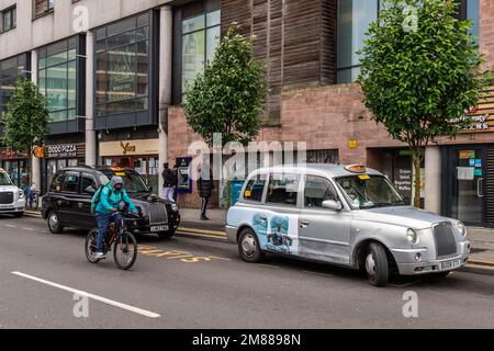 Männlicher Radfahrer von Deliveroo, der in Coventry, West Midlands, Großbritannien, an den gemieteten Taxis vorbeifährt. Stockfoto