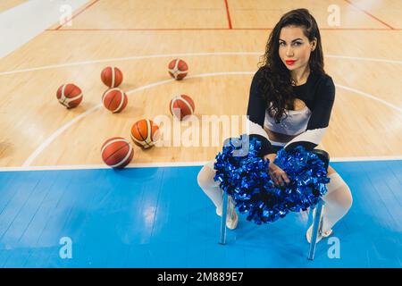 Eine attraktive Cheerleaderin sitzt selbstbewusst und hält ihre Pom-Poms. Sie trägt ein schwarzes Hemd und kniehohe Socken. Ein Gefühl von Macht und Energie. Hinter ihr ist ein Haufen Basketbälle zu sehen. Stockfoto