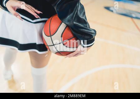 Die untere Hälfte einer Cheerleaderin trägt einen verspielten Minirock, während sie einen Basketball in der Hand hält. Ihre Beine sind fokussiert und zeigen ihren kräftigen und sportlichen Körper. Ihr Gesicht ist nicht zu sehen. Stockfoto