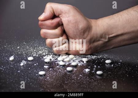 Das Konzept der Suchtbekämpfung. Junge Hand zermalmt weiße Pillen mit der Faust auf grauem Betonboden. Internationaler Tag gegen Drogenmissbrauch. Schließen. Stockfoto