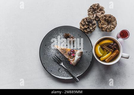 Draufsicht auf einen köstlichen Kuchen auf einem grauen Teller, eine Tasse Tee und Pinienzapfen daneben, Winterästhetik. Hochwertiges Foto Stockfoto