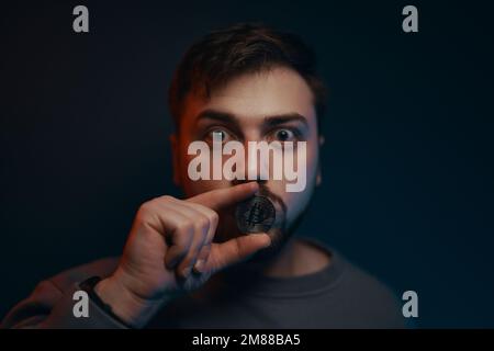 Das Schweigen der Kryptowährung. Ein Mann hält eine Münze in der Nähe seiner Lippen. Stockfoto