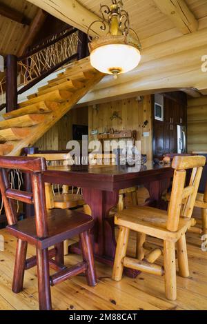 Esstisch aus Zedernholz mit hohen Lehnstühlen im Esszimmer und Blick auf die Treppe aus weißem Kiefernholz im Blockhaus im skandinavischen Stil. Stockfoto