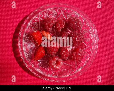 Frische Himbeeren und Erdbeeren Früchte gesunde Snacks. Ein Haufen roter Beeren auf einer verzierten Glasuntertasse auf einem magentafarbenen Tuch Stockfoto
