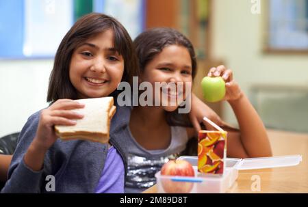 Sie zeigen ihr Mittagessen. Porträt von zwei jungen Schulmädchen, die in der Pause in der Cafeteria zu Mittag essen. Stockfoto