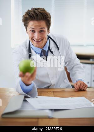 Der einfachste Weg zu einer besseren Gesundheit. Porträt eines positiven jungen Arztes, der einen Apfel hochhält und in die Kamera lächelt. Stockfoto