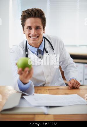 Ich kann genug Gutes darüber sagen. Porträt eines positiven jungen Arztes, der einen Apfel hochhält und in die Kamera lächelt. Stockfoto