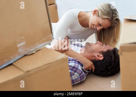 Turteltäubchen in ihrem neuen Zuhause. Ein lächelndes Paar, das kuschelt und flirtet, während es auf dem Boden seines neuen Hauses liegt und von Kisten umgeben ist. Stockfoto