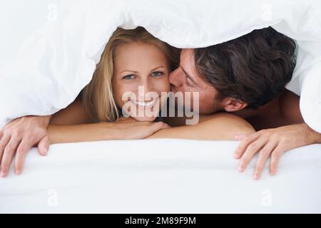 Glücklich mit ihrem Mann. Ein Mann, der eine lächelnde Frau auf die Wange küsst, während er auf einem Bett liegt und unter einem Bettbezug kuschelt. Stockfoto