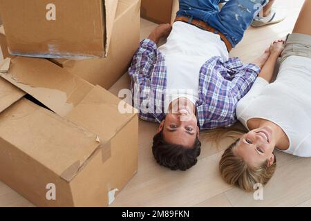 Glücklich in ihrem neuen Zuhause. Porträt glückliches junges Paar, das Händchen hielt, während es in seinem neuen Zuhause auf dem Boden lag und von Kisten umgeben war. Stockfoto