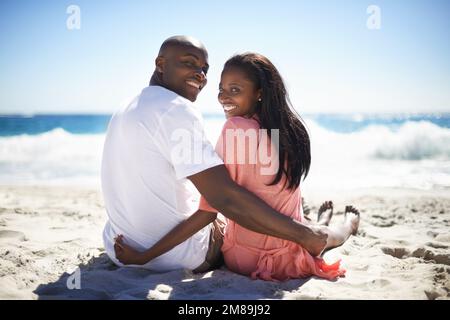 Intimität am Strand. Ein glückliches afroamerikanisches Paar, das über die Schultern blickt, während es am Strand sitzt. Stockfoto