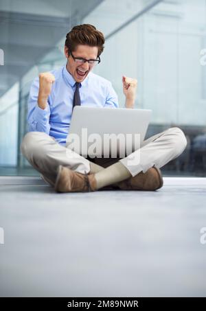 - JA, ICH WEISS Ein junger Geschäftsmann, der auf dem Boden seines leeren Büros sitzt und mit einem Laptop auf dem Schoß feiert. Stockfoto