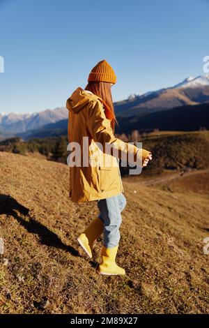 Die Frau geht auf einem Hügel in voller Länge spazieren und sieht die Berge in einem gelben Regenmantel und Jeans im Herbst auf einer schönen Reise bei Sonnenuntergang wandern Stockfoto