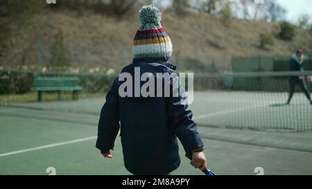 Das Kind spielt in der Wintersaison mit seinen Eltern Tennis. Ein Kind trägt einen Mantel und schlägt mit einer Beanie auf den Ball. Der kleine Junge spielt draußen Sport Stockfoto