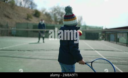 Das Kind spielt in der Wintersaison mit seinen Eltern Tennis. Ein Kind trägt einen Mantel und schlägt mit einer Beanie auf den Ball. Der kleine Junge spielt draußen Sport Stockfoto