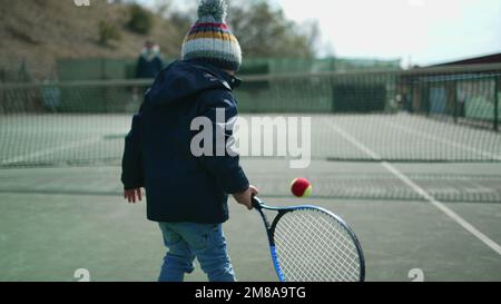 Das Kind spielt in der Wintersaison mit seinen Eltern Tennis. Ein Kind trägt einen Mantel und schlägt mit einer Beanie auf den Ball. Der kleine Junge spielt draußen Sport Stockfoto