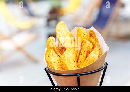 Im Ofen gebackene Kartoffelsplitter mit Meersalz und Kräutern. Pommes Frites in einer recycelbaren Papiertüte auf einem verschwommenen Straßenhintergrund, beliebtes Fast Street Food Stockfoto