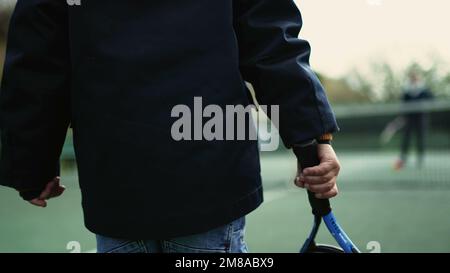 Das Kind spielt in der Wintersaison mit seinen Eltern Tennis. Ein Kind trägt einen Mantel und schlägt mit einer Beanie auf den Ball. Der kleine Junge spielt draußen Sport Stockfoto