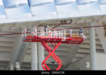Bauarbeiter stehen in der Schere und heben Kranschaufel an, während sie auf hoher Höhe auf der Baustelle arbeiten Stockfoto