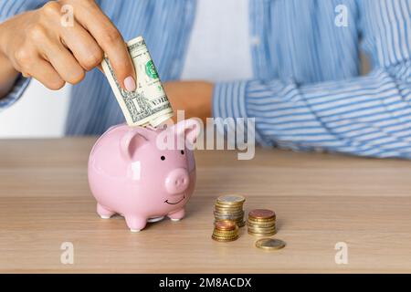 Nahaufnahme der Hände einer Frau Legen Sie Dollar-Papiergeld in ein Sparschwein, neben einem Rechner auf dem Tisch.Sparen Sie Geld in einer Krise und Inflation Stockfoto
