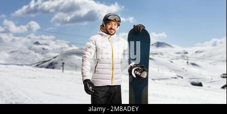 Ein junger Mann steht auf einem Berg mit einem Snowboard und schaut in die Kamera Stockfoto