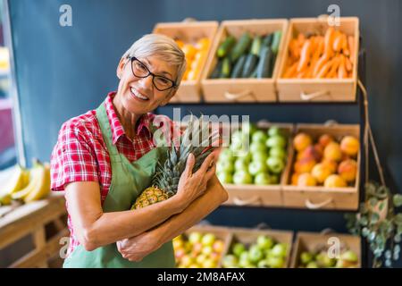 Reife Frau arbeitet im Obst- und Gemüseladen. Sie hat Ananas in der Hand. Stockfoto