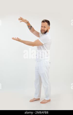 Porträt eines lächelnden, gutaussehenden barfuß-alten Mannes mittleren Alters, der auf weißem Hintergrund auf dem Boden steht, die Hände hebt und zeigt. Stockfoto